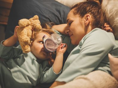 Two year old girl drinking water from the bottle in bed, holding Teddy bear and lying down next to her mother.
