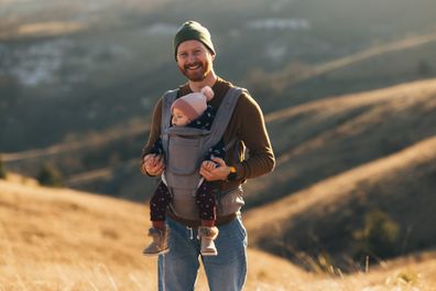 Smiling dad wearing a beanie carries baby in a sling on a hike during golden hour with hills in background.
