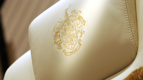 A gold-threaded, embroidered Trump family crest on the headrest of a chair.