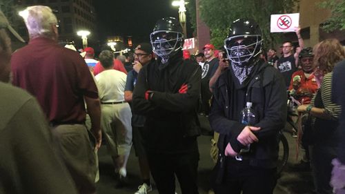 Black clothed protestors - appearing to be members of Antifa - watch on as Trump supporters leave the rally. (9NEWS/Lizzie Pearl)