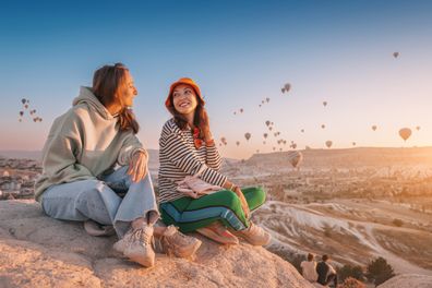 Girl friends sitting on a clifftop viewpoint and admiring view of majestic flying hot air balloons in Cappadocia