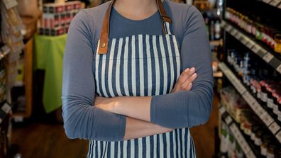 Confident female business owner of a supermarket standing between shelves while facing camera smiling with arms crossed