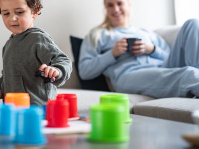 Mum sits on a lounge watching her toddler play with toys.