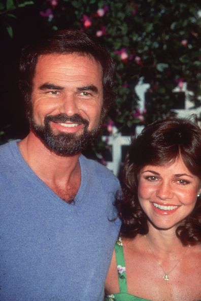 American actor Burt Reynolds smiles with his girlfriend, actor Sally Field, while attending an outdoor event. He wears a blue V-neck sweater, and has a beard and mustache. 