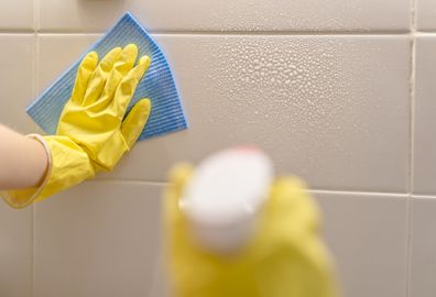 woman washes ceramic tiles on kitchen wall with detergent. Female holding a cleaning spray and a sponge in her hands. Woman's hand in yellow rubber protective glove with rag cleaning tiles. 
