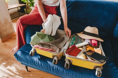 Beautiful smiling young woman packing personal belongings in a suitcase for travelling looking at list at tablet sitting on the sofa in the living room.