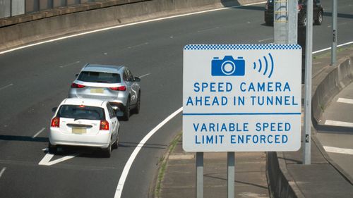 Cars with New South Wales number plates travelling on the Eastern Distributor tollway that skirts the central business district of Sydney. They are passing a sign warning of speed cameras in the approaching tunnel.  One of the cars is tailgating the car in front.  Another car is obscured behind a pillar on the right. This image was taken from a pedestrian walkway linking Woolloomooloo Bay and The Domain near the Art Gallery of NSW on a hot and sunny afternoon on 1 March 2025.