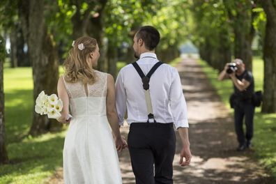 Bride holding bouquet of flowers and walking with groom on single track in park, photographer taking photo with digital camera seen in background.