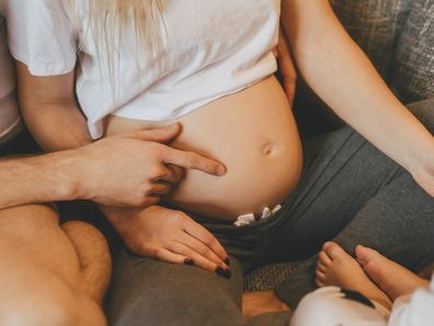 Stylish modern pregnant mother, father and son are spending time together at home. Male hand shows on tummy. Pregnancy, family, parenthood, preparation and expectation concepts. Warm colors.