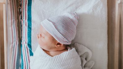 A baby just born at the hospital rests in a hospital bassinet crib, wrapped in a swaddle and wearing a beanie hat.