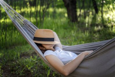 Woman resting in a hammock in a summer garden covering her face with a straw hat. Summer vacation concept