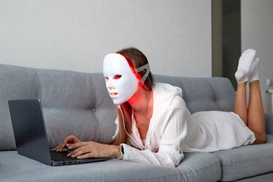 A beautiful girl with an LED mask on her head works at home on a laptop. Home skin care concept.