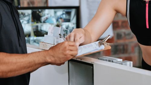 Asian man signing up for gym membership with caucasian trainer. Young coach behind reception pointing and showing where to sign. Two active and fit people standing together in health and fitness club