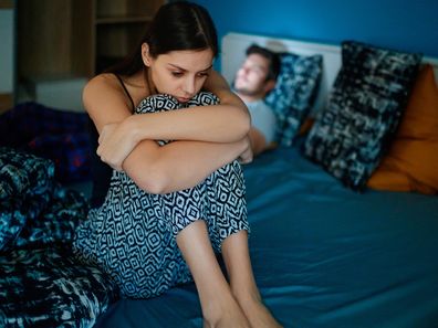 Stock image of a woman sitting on a bed's edge while her partner is lying in bed in the background.