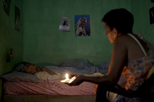 A woman sits beside her sick mother holding a candle. She says the hours of intermittent electricity supply in Venezuela affects her capacity to care for, bathe and feed her mother.