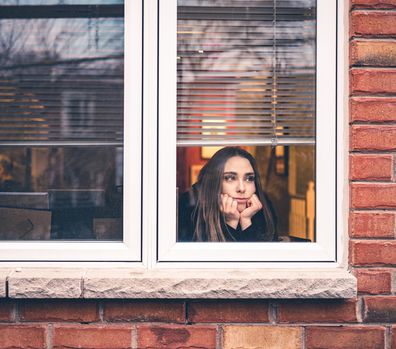 Young woman in lockdown, sitting inside her home, close to the window looking out. She is practicing social distancing due to COVID-19