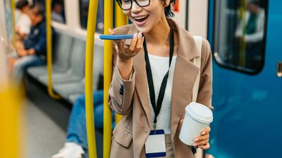 Beautiful young Japanese Asian businesswoman sending a voice message using her phone and drinking coffee to go while riding in a subway train in Sydney in Australia.