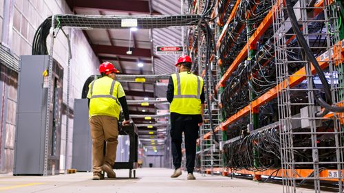 Two technicians wearing PPE walking past racks of equipment in a data center.