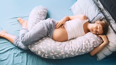 Beautiful pregnant woman relaxing or sleeping with belly support pillow in bed. Young mother waiting of a baby. Concept of pregnancy, maternity, healthcare, gynecology, medicine.