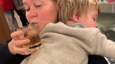 Comedian Amy Schumer attempting to take a sip of her drink while holding her sleeping child