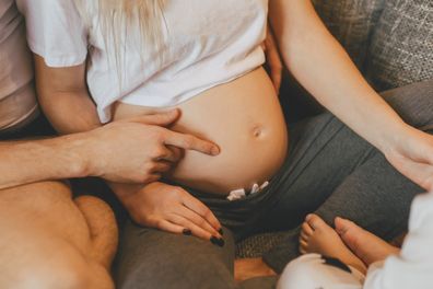 Stylish modern pregnant mother, father and son are spending time together at home. Male hand shows on tummy. Pregnancy, family, parenthood, preparation and expectation concepts. Warm colors.
