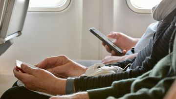 Long shot across laps of older person hands using cell phone.  Framed by seat back and clipped airplane windows.  