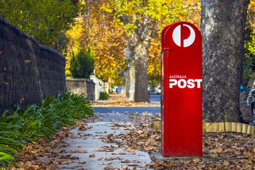 Adelaide, Australia - May 5, 2021: full length view of Australia Post Red Street Posting Box in quiet, leafy established suburban street. Autumn colours, brush fence, leaf litter, pavement, green plants