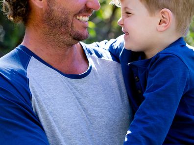 Father holding young son outdoors in sunlight in park - looking away from camera