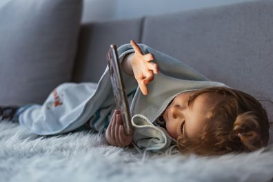 Close up of adorable kid laying down on the bed by holding smartphone at night. Photo of Little kid in bed under a blanket looking at the smartphone at night. Happy smiling baby boy laying on his tummy on the bed under a blanket and plays on a smartphone in a game in the dark. The child's face is illuminated by a bright monitor.