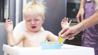 Cranky baby in the early morning, sitting on a baby chair in the kitchen. Being fed baby food from a spoon.