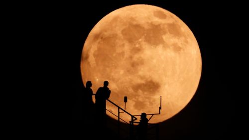 People are silhouetted as an almost full moon rises above the sky.