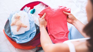 Pregnant woman packing suitcase, bag for maternity hospital at home, getting ready for newborn birth, labor. Pile of baby clothes, necessities and pregnant women at awaiting.