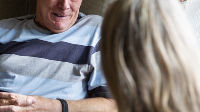 Nurse conversing with a senior patient in the North East of England while he is sitting in a chair in his living room.