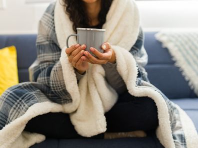 Close up of a young woman wrapped in a quilt relaxing on the sofa during autumn weather at home drinking hot tea or chocolate