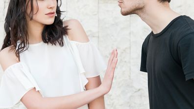 A young guy is leaning in to kiss a girl be she is frowning and making a hand gesture to stop him. They are wearing casual clothes.