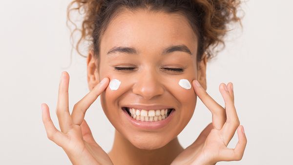 Skin care. Laughing girl applying moisturising cream on her face over white background