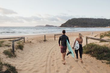 A rear-view shot of a young man and woman holding a surfing board on a beach in Sydney, Australia.