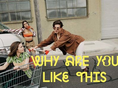 Poster for show "Why are you like this." Man in centre is pushing a trolley with a woman in it , while another woman leans against a car in the background.