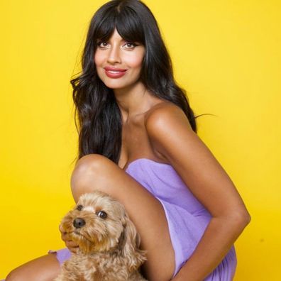Jameela Jamil wears a purple lilac dress on a yellow background.  She is in a crouching position, petting a small brown dog that also appears in the photo.