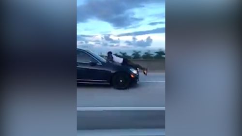 The man can be seen on the bonnet of the car which is travelling fast along the highway.