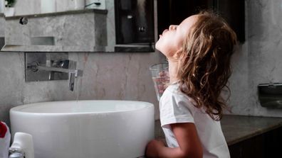 Cute little curly child girl rinses her mouth with water, looking at mirror and spits water into the sink, at domestic bathroom, side view.