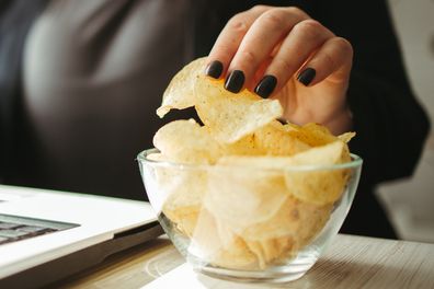 Compulsive overeating, mindless snacking, junk food, unhealthy meals. Woman eating chips from bowl at her workplace