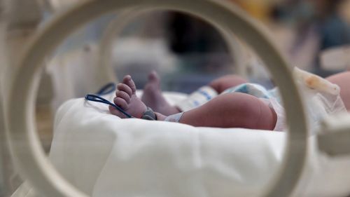 A baby's feet are visible in a hospital incubator. The baby is wrapped in a blanket and has a blue strap around its foot. The baby is in a hospital setting, and the image conveys a sense of care