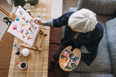 From above photo of senior woman painting an art canvas while sitting on a couch in a living room.