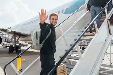 A young backpacker boards a plane on a budget trip. He walks up the steps to the plane with his suitcase and smiles at the camera while waving.