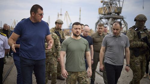 In this photo provided by the Ukrainian Presidential Press Office, Ukrainian President vZelenskyy, center, surrounded by ambassadors of different countries and UN officials, visits a port in Chornomork during loading of grain on a Turkish ship, background, close to Odesa, Ukraine, Friday, July 29, 2022. (Ukrainian Presidential Press Office via AP)