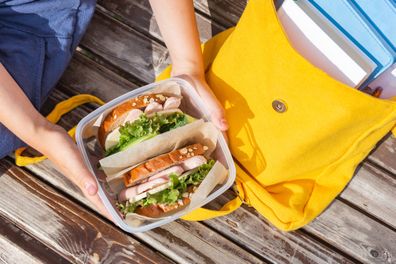 Lunch box in the hands of a child. Sandwiches with bread, lettuce, cucumber and sausages in a plastic container. Snack, school breakfast, lunch. Back to school. Lunch break. Yellow backpack with books