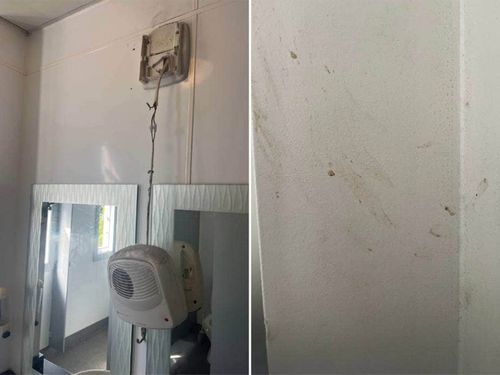 A bathroom heater hangs by an electrical wire, while a wall is smeared with unknown matter.