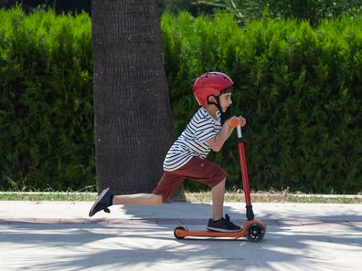 Portrait of little boy riding kick scooter on sidewalk in outdoor. He is wearing a red and white striped t-shirt and a blue short. The scooter and helmet are red. The background is a concrete high wall. Shot in outdoor daylight with a full frame mirrorless camera.