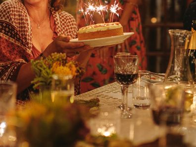 Young woman celebrating her birthday with her friends. Female holding birthday cake sitting with friends around dining table outdoors.